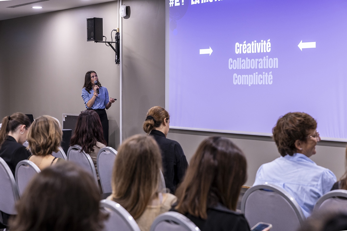 Une femme, vêtue d'une chemise bleue, s'adresse à un public à l'aide d'un micro et d'un pointeur. Elle est positionnée à gauche, près d'un écran de projection violet affichant des mots clés en français : "Créativité," "Collaboration," et "Complicité." Un groupe de participants, principalement des femmes, est assis face à elle, écoutant attentivement. L'ambiance est professionnelle et studieuse.